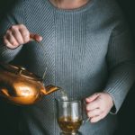 woman pouring tea from antique copper kettle into a glass mug in front of a table