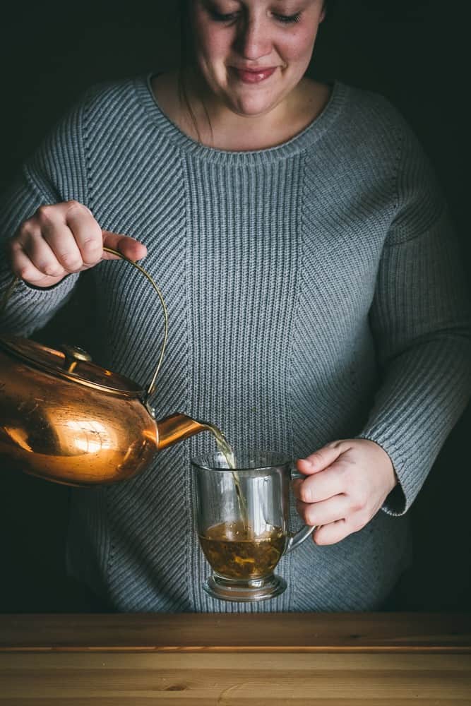 woman pouring tea from antique copper kettle into a glass mug in front of a table