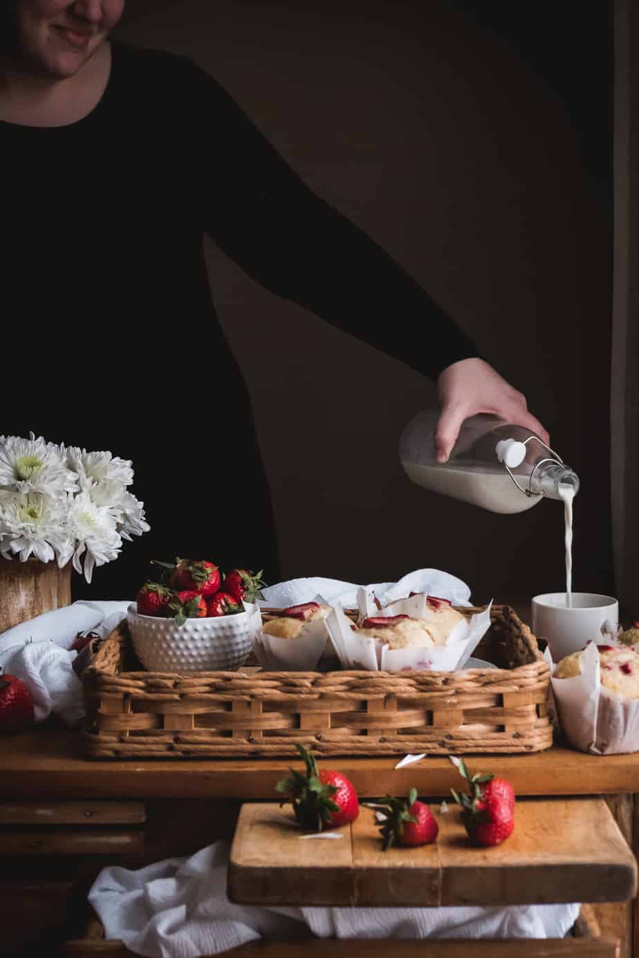 woman pouring milk into cup next to tray of muffins and strawberries